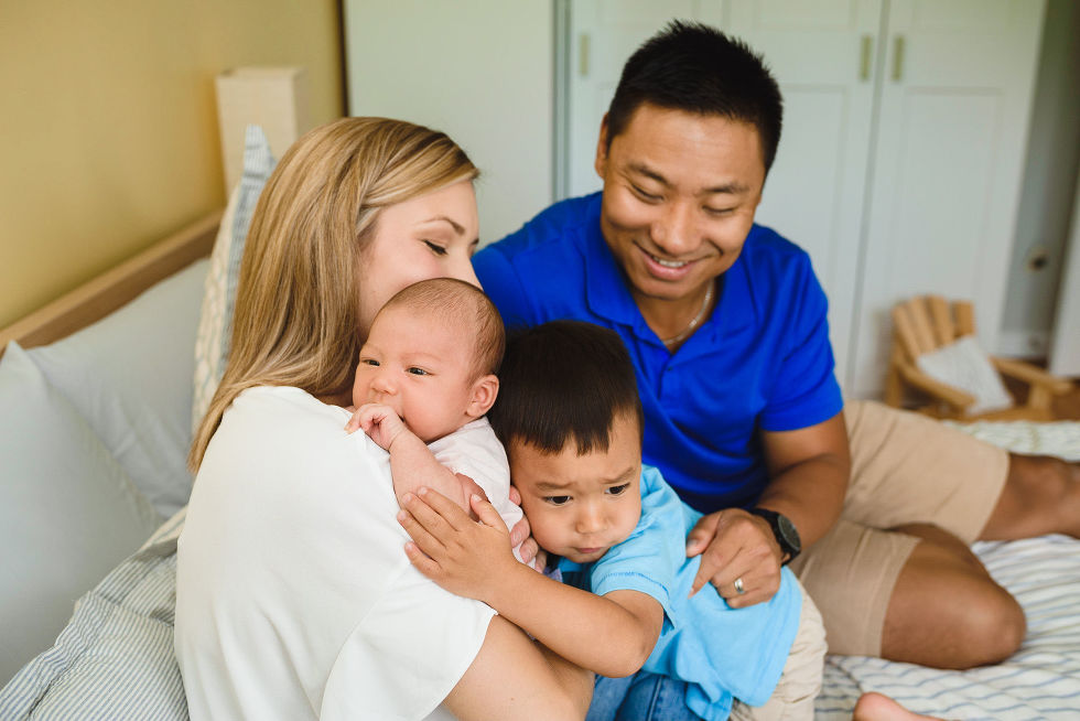 A mother holding her newborn baby as her toddler and husband wrap their arms around her on their bed Toronto family photography