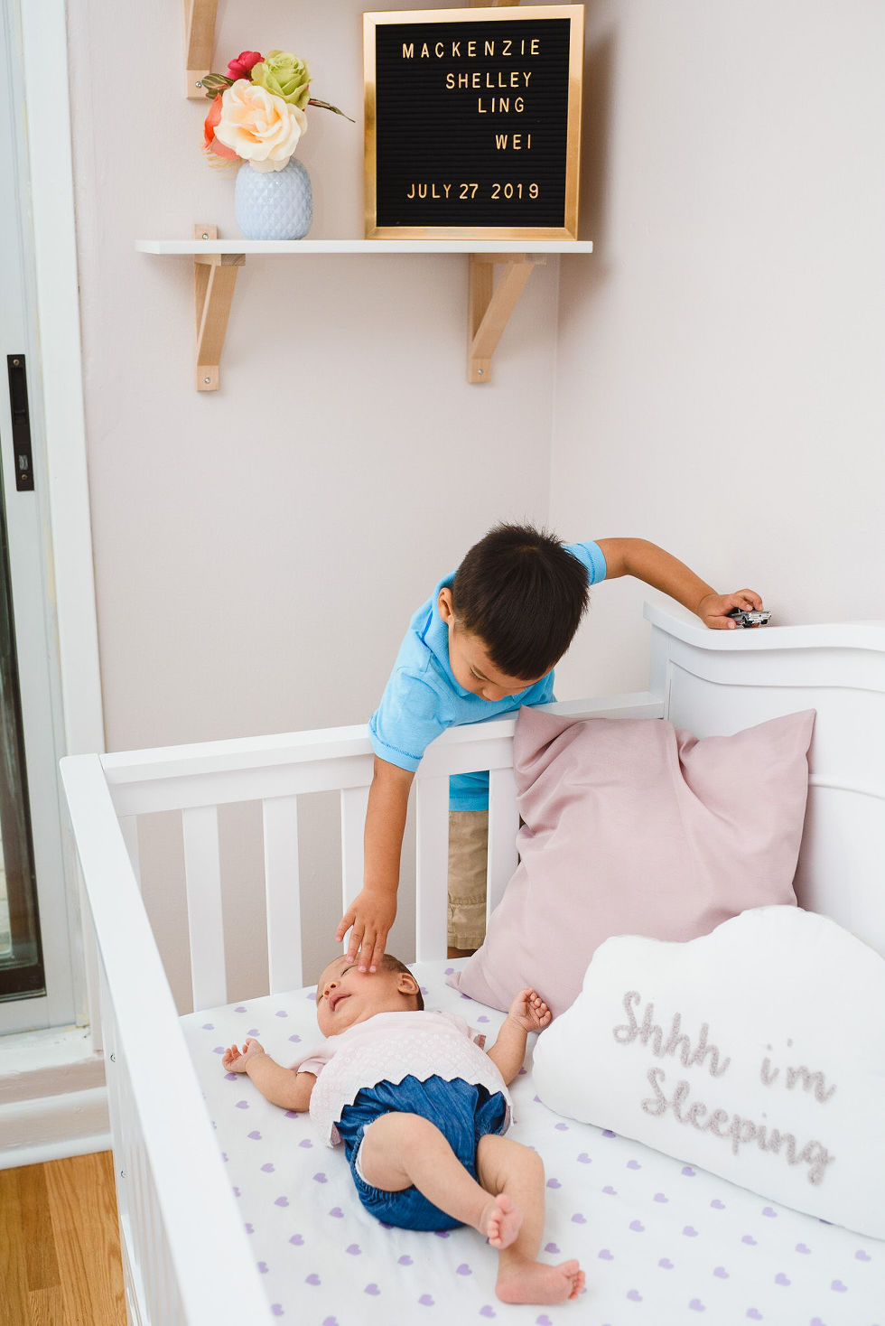 A toddler reaching into a crib to say hi to his baby sibling with a frame above the crib with a birth announcement on it Toronto family photography